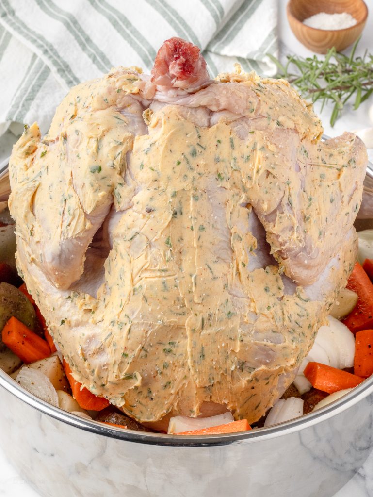 A whole chicken sitting on a bundt pan overtop of potatoes, carrots, and onions. The chicken is slathered with a garlic herb butter and ready to go in the oven and roast.