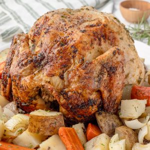 Herb Roasted Chicken resting upon roasted potatoes, carrots, and onions. The chicken is juicy, the skin is golden brown and crispy with speckling of herbs.