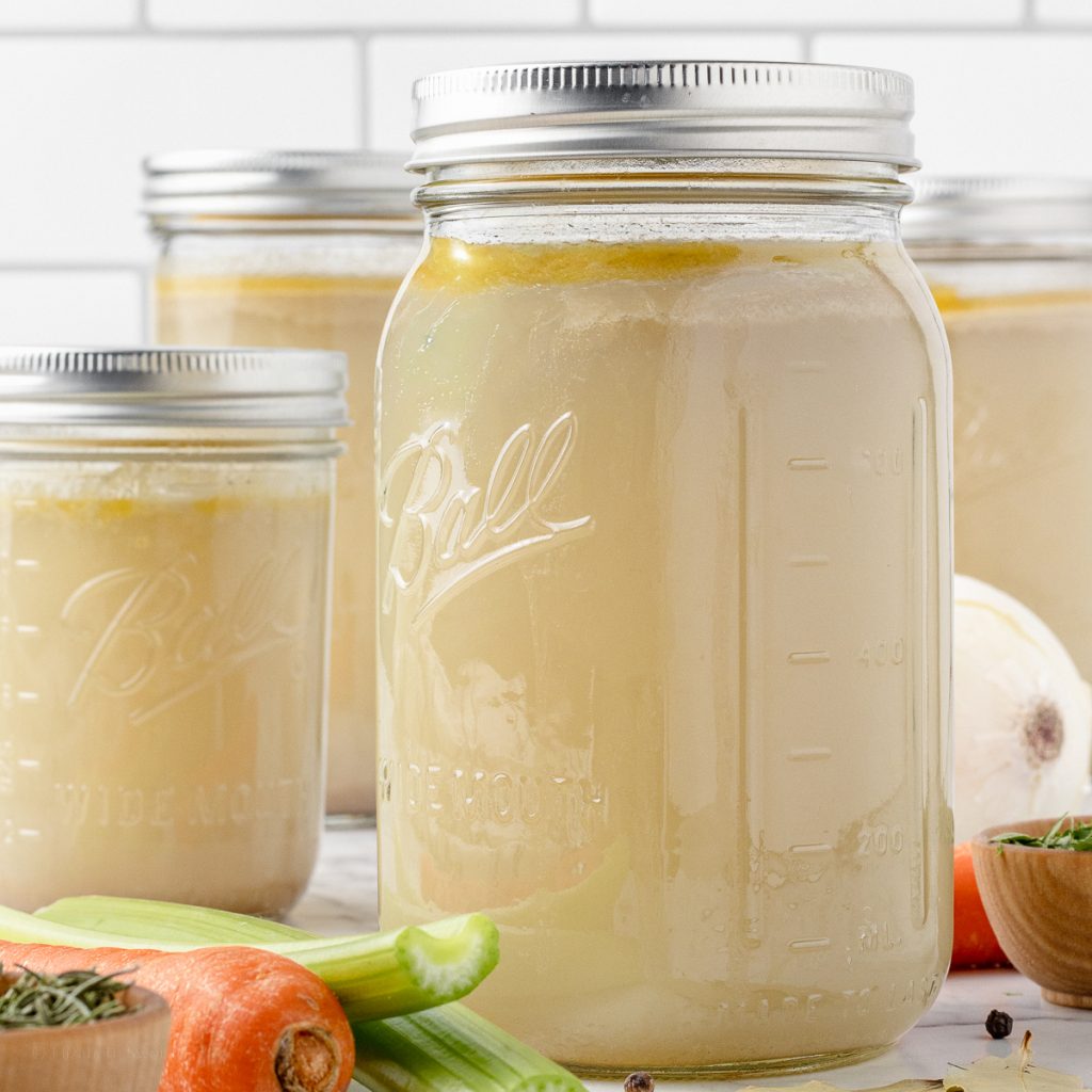 4 jars of Homemade Chicken Stock surrounded by carrots, celery, onions, herbs, and spices.