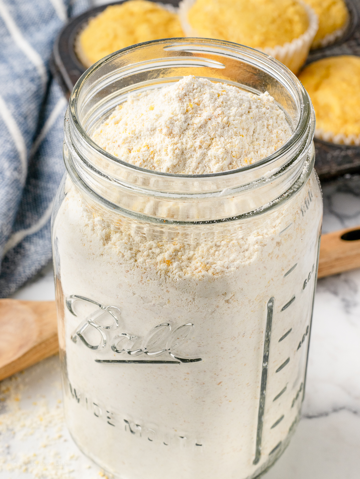 Homemade, self rising Cornbread Mix in a jar with sweet cornbread muffins in the background.