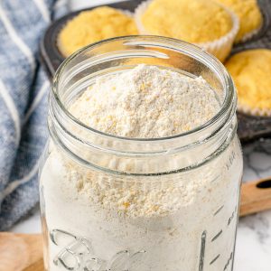 Close up of homemade, self rising Cornbread Mix in a jar with sweet cornbread muffins in the background.