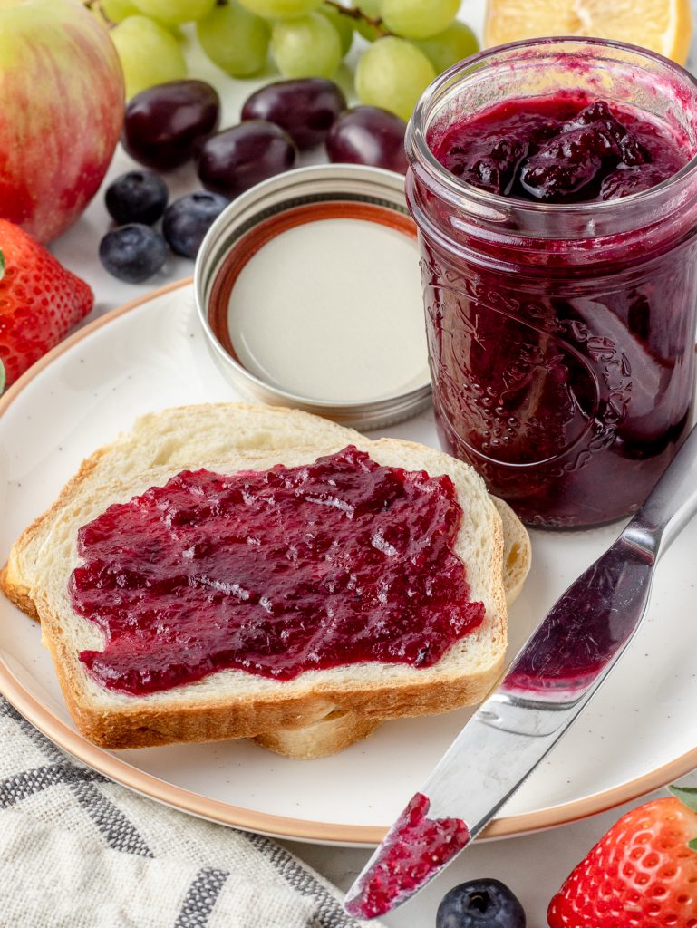 Plate with a jar of jam and homemade sandwich bread slathered with the Mixed Fruit Jam. The knife is on the side for scooping out more.