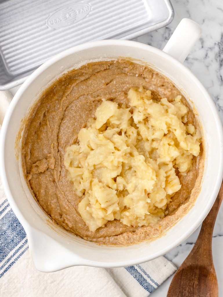 Banana bread batter with mashed banana sitting on top ready to stir in. There is a large spoon and bread pan on the side.