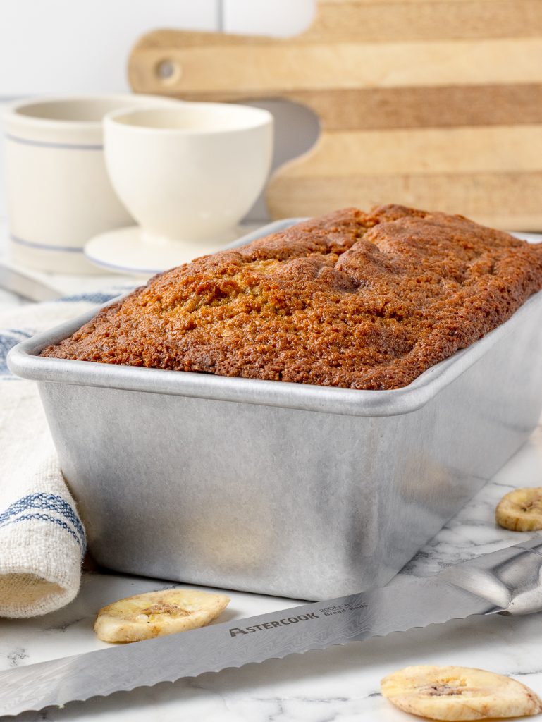 Baked bread in a bread pan with serrated knife and butter bell on the side.