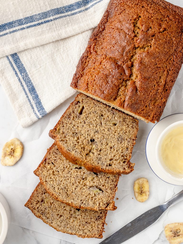 Overhead image of banana bread with 3 slices of the bread laying down. There is a knife and butter bell filled with butter on the side.
