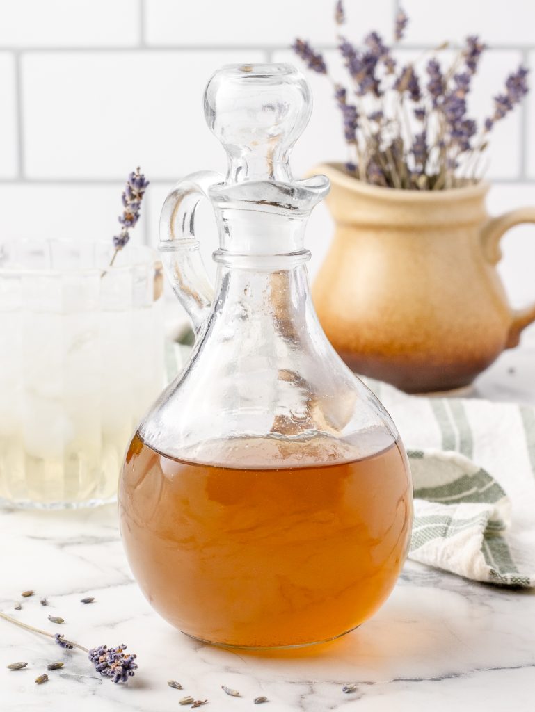 Bottle of the Lavender Simple Syrup with a drink made from it in the background. There is also a vase of dried lavender and lavender buds in the picture.