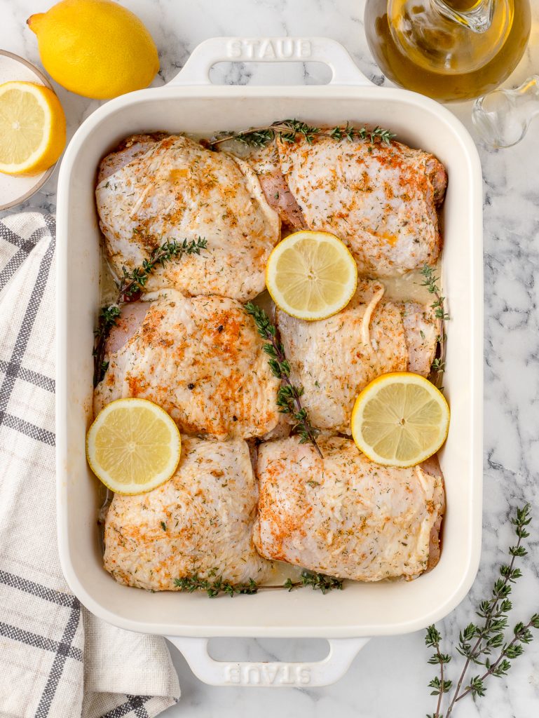 The bone in, skin on chicken thighs coated with olive oil and spices, sitting in a lemon juice mixture. They are surrounded by lemons and fresh thyme, ready to go in the oven.