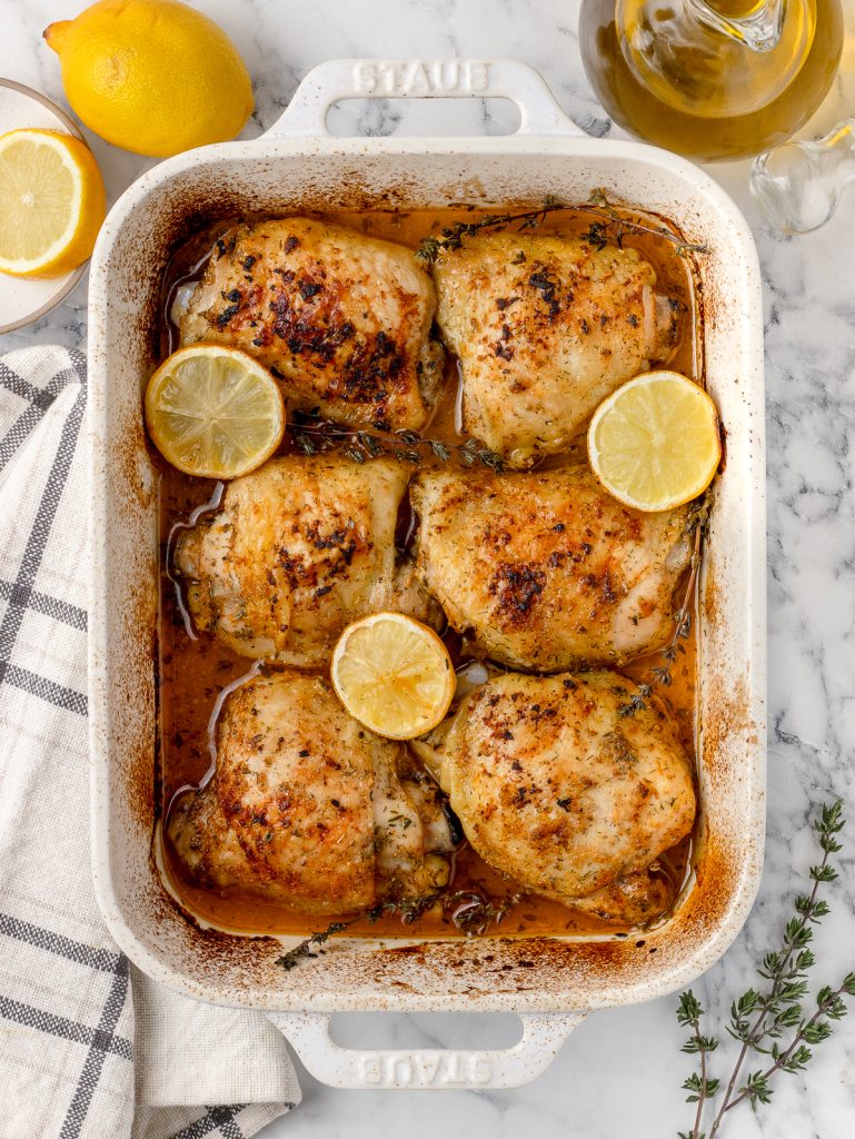 Baked chicken thighs straight out of the oven. They have a crispy skin and are surrounded by roasted lemons and thyme.