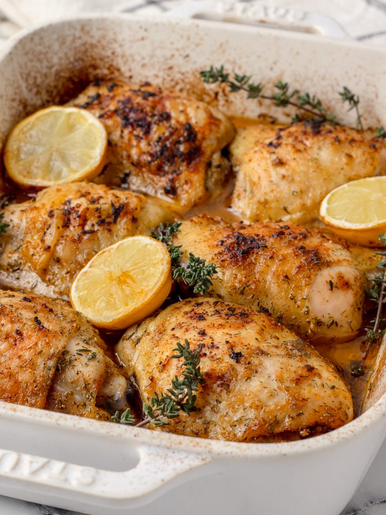 Baked Lemon Chicken Thighs in a ceramic dish with roasted lemons and thyme.