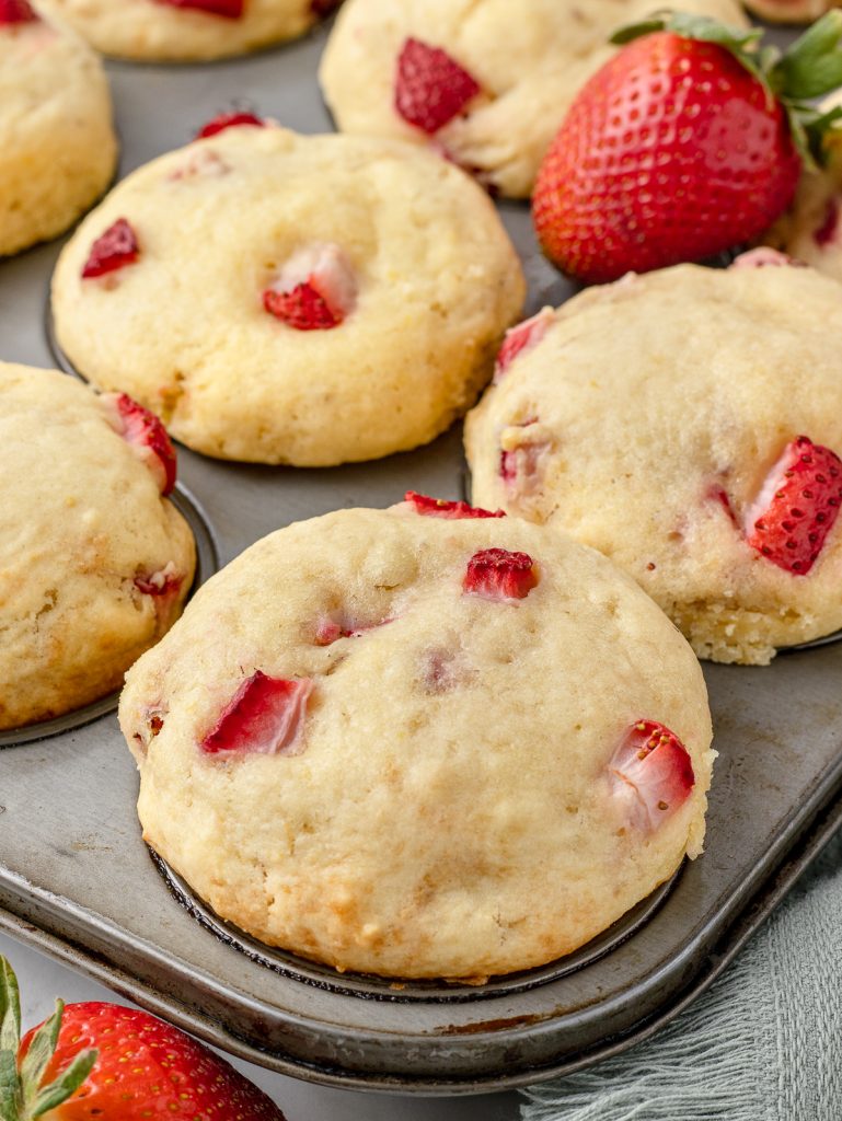 Lemon Strawberry Muffins in a muffin tin. You can see the strawberries and flecks of lemon zest. They are tall with perfectly rounded dome tops.