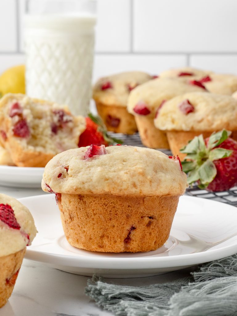 Muffin sitting on a plate ready to eat. There is one in the background that is half eaten, and a cooling rack with more muffins. There is a glass of milk too.