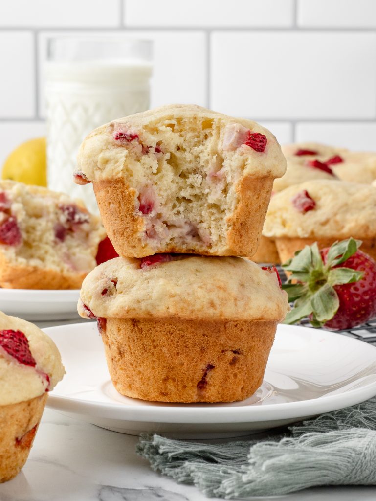 Stack of Lemon Strawberry Muffins. The top one has a bite taken from it and you can see the soft, tender insides with chunks of strawberries and flecks of lemon zest. There are more muffins and a glass of milk in the background.