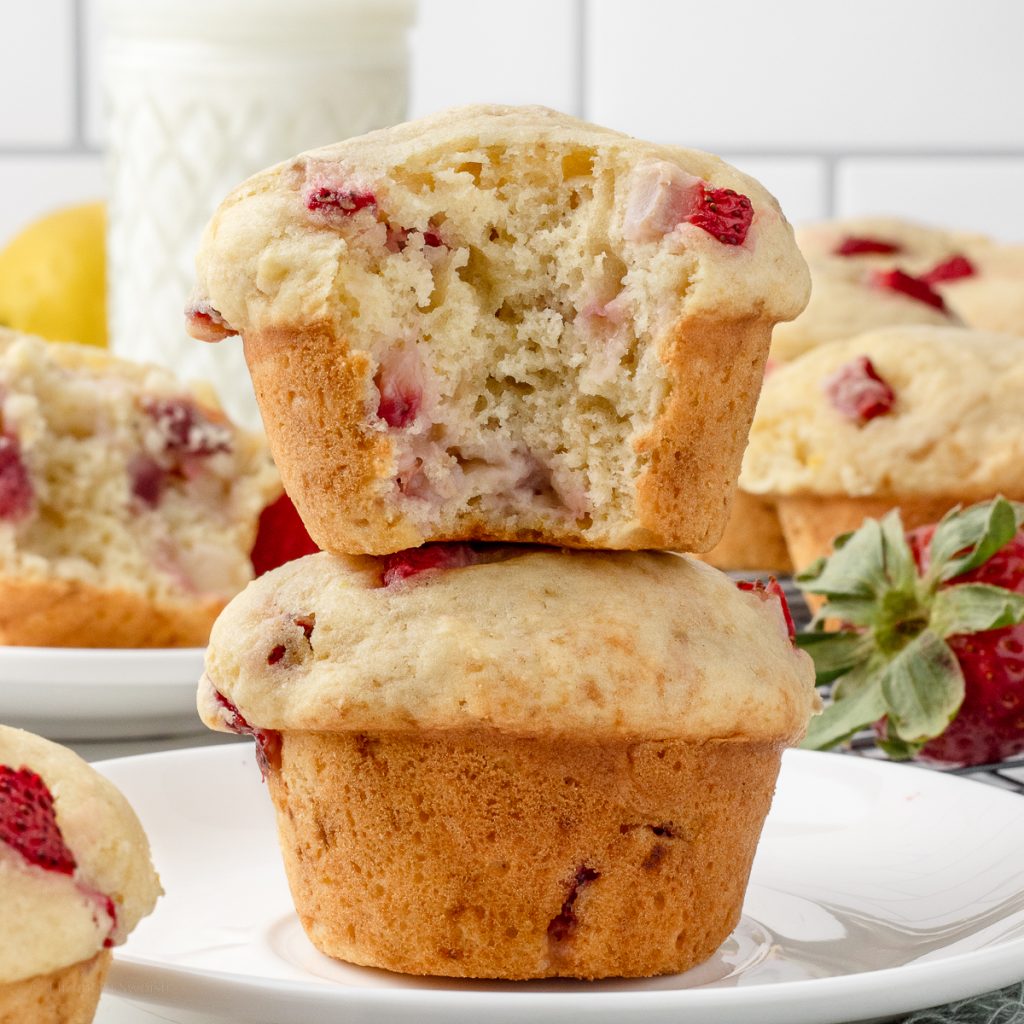 Stack of Lemon Strawberry Muffins. The top one has a bite taken from it and you can see the soft, tender insides with chunks of strawberries and flecks of lemon zest.