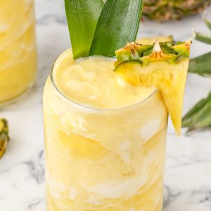 Pina Colada Mocktail in a glass and garnished with a pineapple wedge and a couple pineapple leaves. The mocktail is a pretty yellow with swirls of white from the coconut cream.