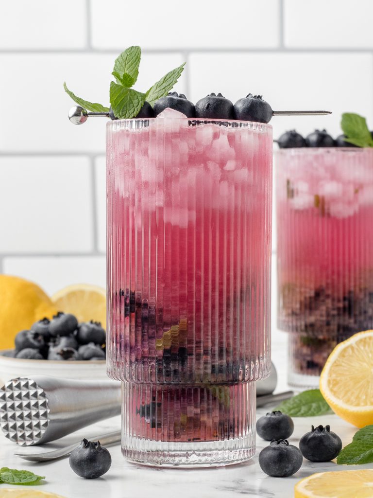 Close up of the magenta mocktail to see the muddled blueberries and mint on the bottom. It is garnished with more fresh mint and fresh blueberries.