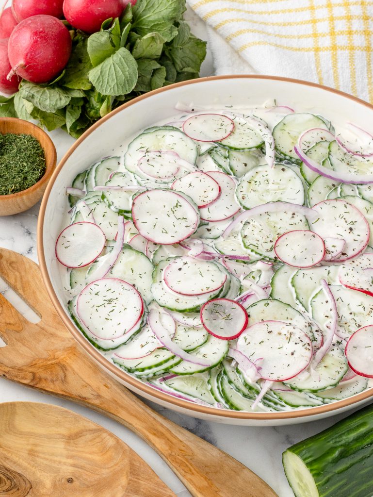 Cucumber Radish Salad tossed in a sweet and creamy dressing with red onion, dill, and black pepper. Serving spoons and additional dill, cucumber, and radish are on the side.