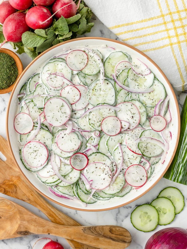 Overhead image of the salad in a big serving bowl. The finely shaved cucumbers, radishes, and onions are tossed in a creamy dressing and topped with dill and black pepper.