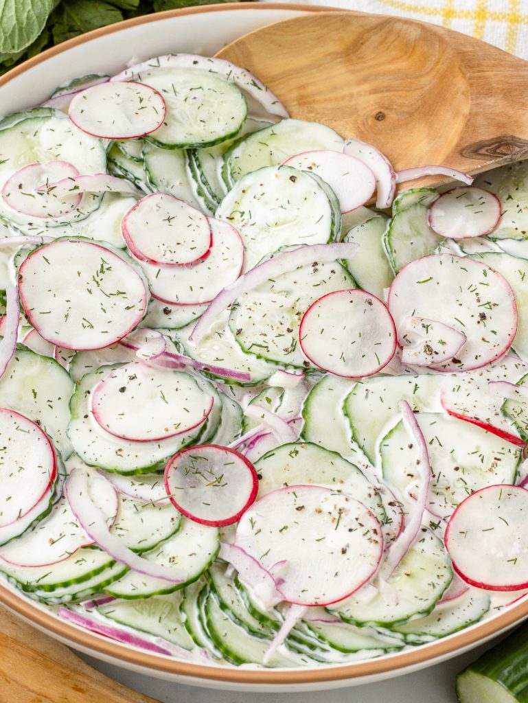 The salad in a bowl. You can see the finely shaved cucumbers, radishes, and onions are tossed in a creamy dressing and topped with dill and black pepper.