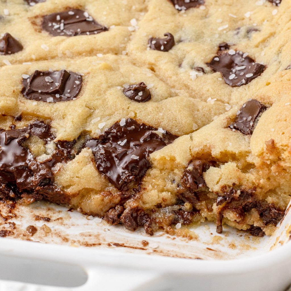 Baked Scoopable Cookies topped with flaky sea salt in a baking dish. There is some removed so you can see the gooey, chocolatey, texture of it.