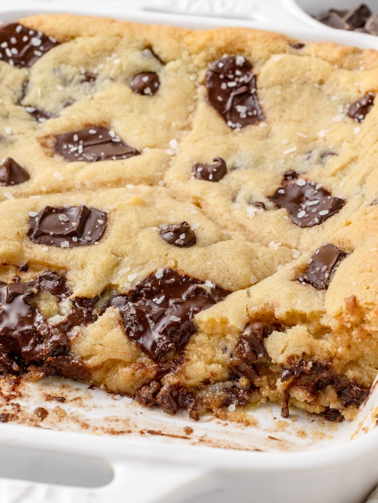 Baked Scoopable Cookies topped with flaky sea salt in a baking dish. There is some removed so you can see the gooey, chocolatey, texture of it. There is more chocolate chips and chunks in the background.
