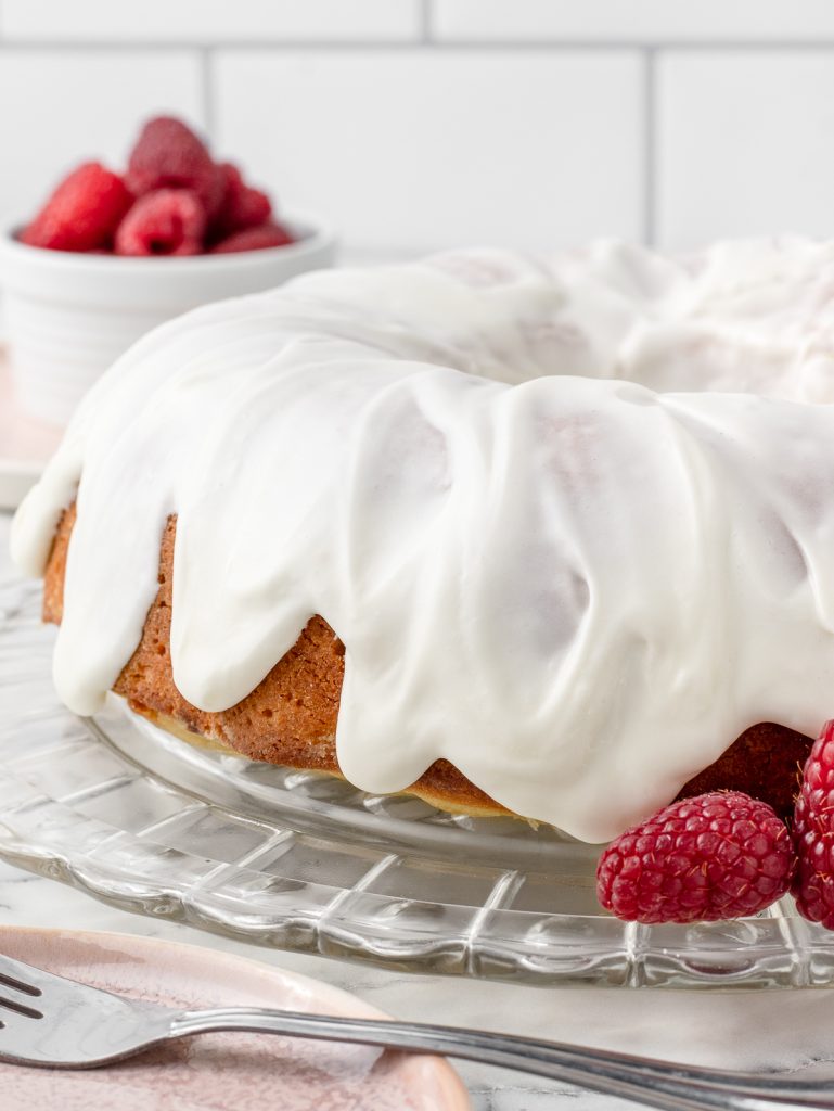 Raspberry White Chocolate Bundt Cake glazed with a thick but light sweet cream glaze. There are fresh raspberries, plates, and forks on the side for serving.