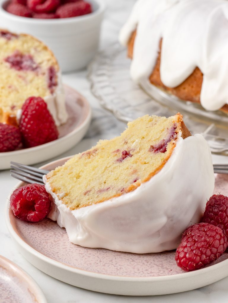 A slice of bundt cake on a plate with additional fresh raspberries on the side. There is another slice of cake on a plate in the background as well as the whole cake.