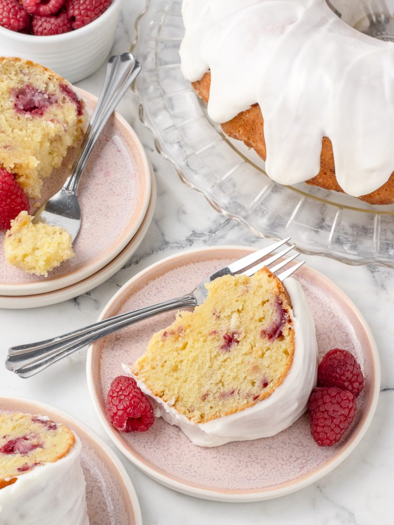3 plates of White Chocolate Raspberry Bundt Cake with fresh raspberries on the side. The big bundt cake is in the background ready for more slices to be sliced.