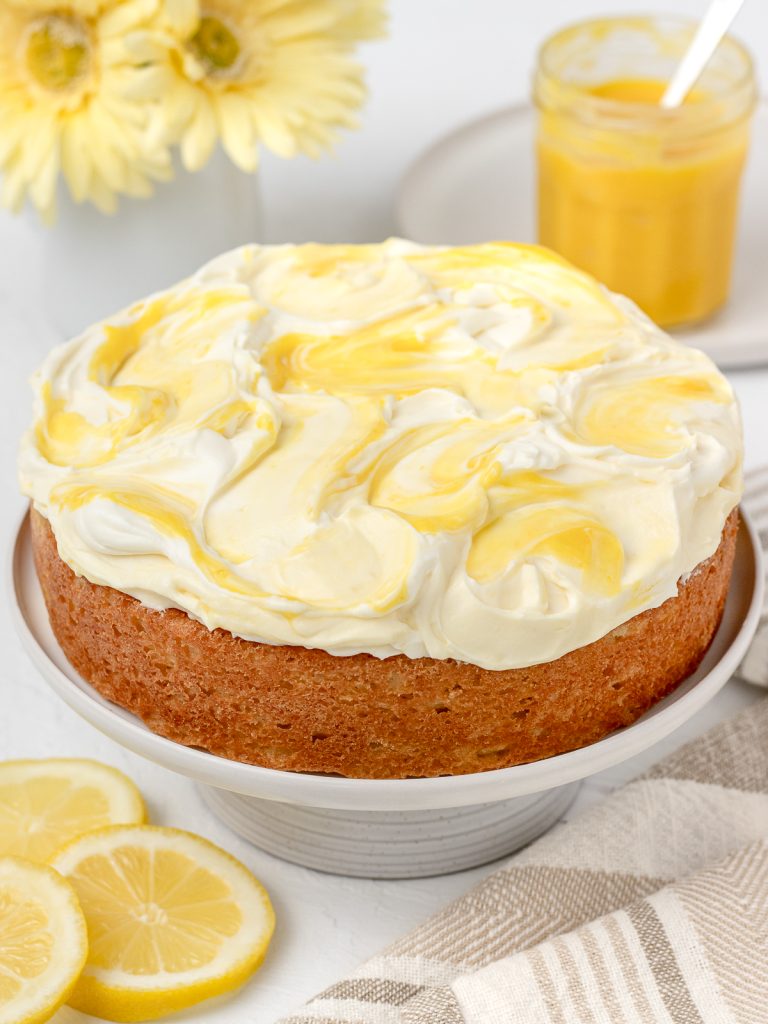 Lemon Curd Cake sitting on a cake stand with more lemon curd and slices of lemon on the side. The frosting on top of the cake is beautifully swirled with lemon curd.