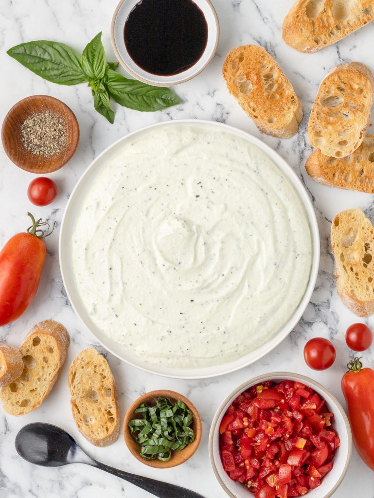 Plate of creamy garlic basil ricotta. On the side are marinated tomatoes, basil, balsamic glaze, fresh cracked black pepper, and crostinis.