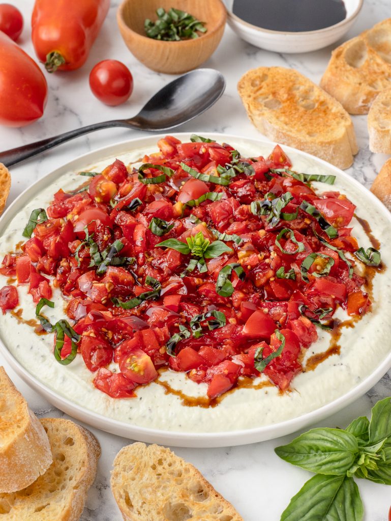 Big plate of Bruschetta Dip with crostinis on the side and a spoon for serving.
