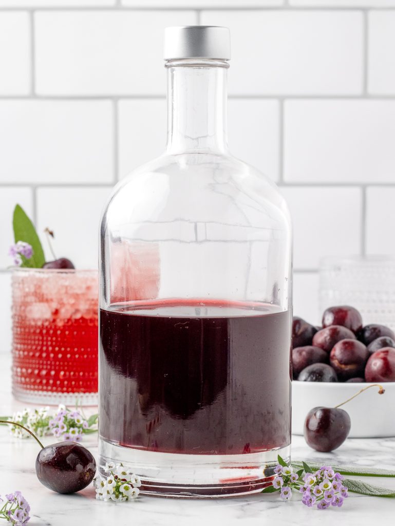 The shrub in a glass bottle with a prepared drink and fresh cherries in the background.