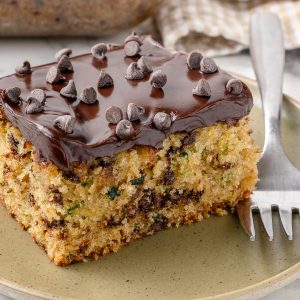 Slice of Chocolate Chip Zucchini Cake with a fork on the side for eating. It's a soft crumb with flecks of zucchini and chocolate chips, with a super creamy chocolate ganache frosting topped with mini chocolate chips.