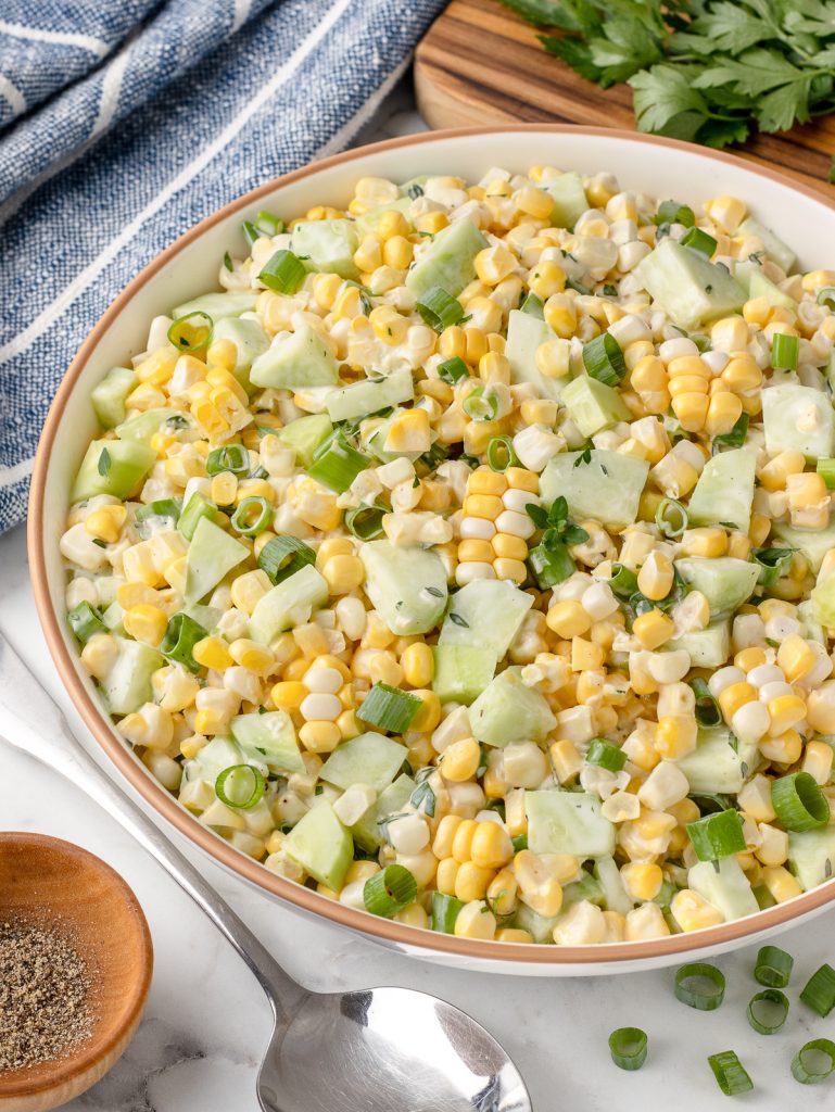 A bowl of this yummy summer salad. It's loaded with raw sweet corn, cucumbers, green onion, and a creamy fresh herb dressing. There is a spoon and black pepper on the side for serving.