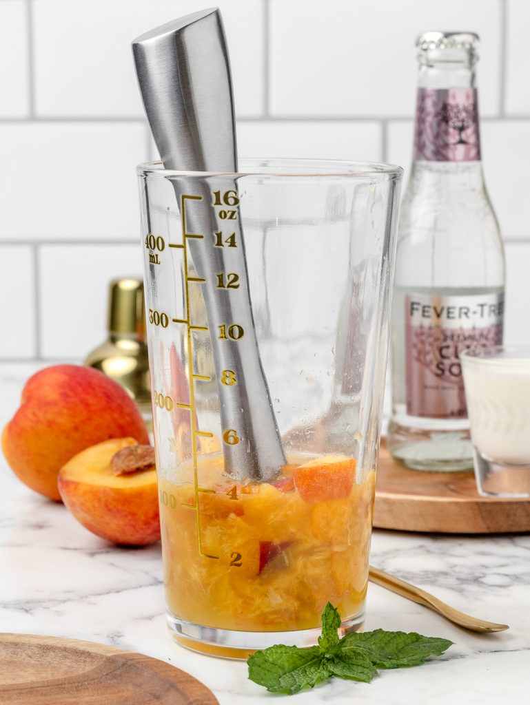 Large glass with a muddler and the muddled fresh peaches and peach syrup.